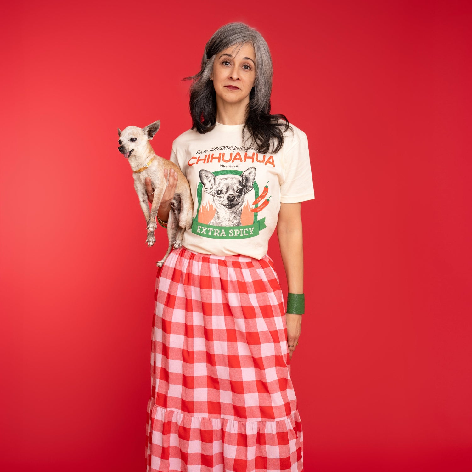 Woman wearing a chihuahua tee shirt and holding an elderly chihuahua against a red background