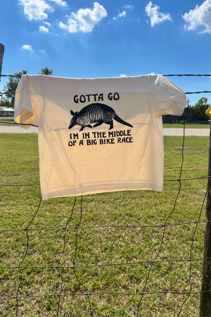 An ivory boxy style t-shirt with a hand printed armadillo graphic, hanging on a rustic barbed wire fence in front of a blue sky