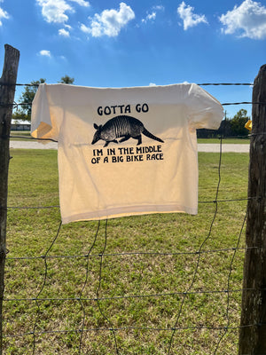 An ivory boxy style t-shirt with a hand printed armadillo graphic, hanging on a rustic barbed wire fence in front of a blue sky
