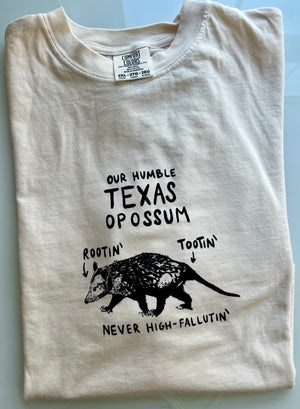 Folded ivory tshirt with a hand drawn possum that says: Our Humble Texas Opossum: Rootin' Tootin' Never High Fallutin