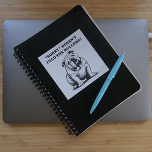 A black notebook with a sticker that says Sorry Doesn't Feed the Bulldog in black and white