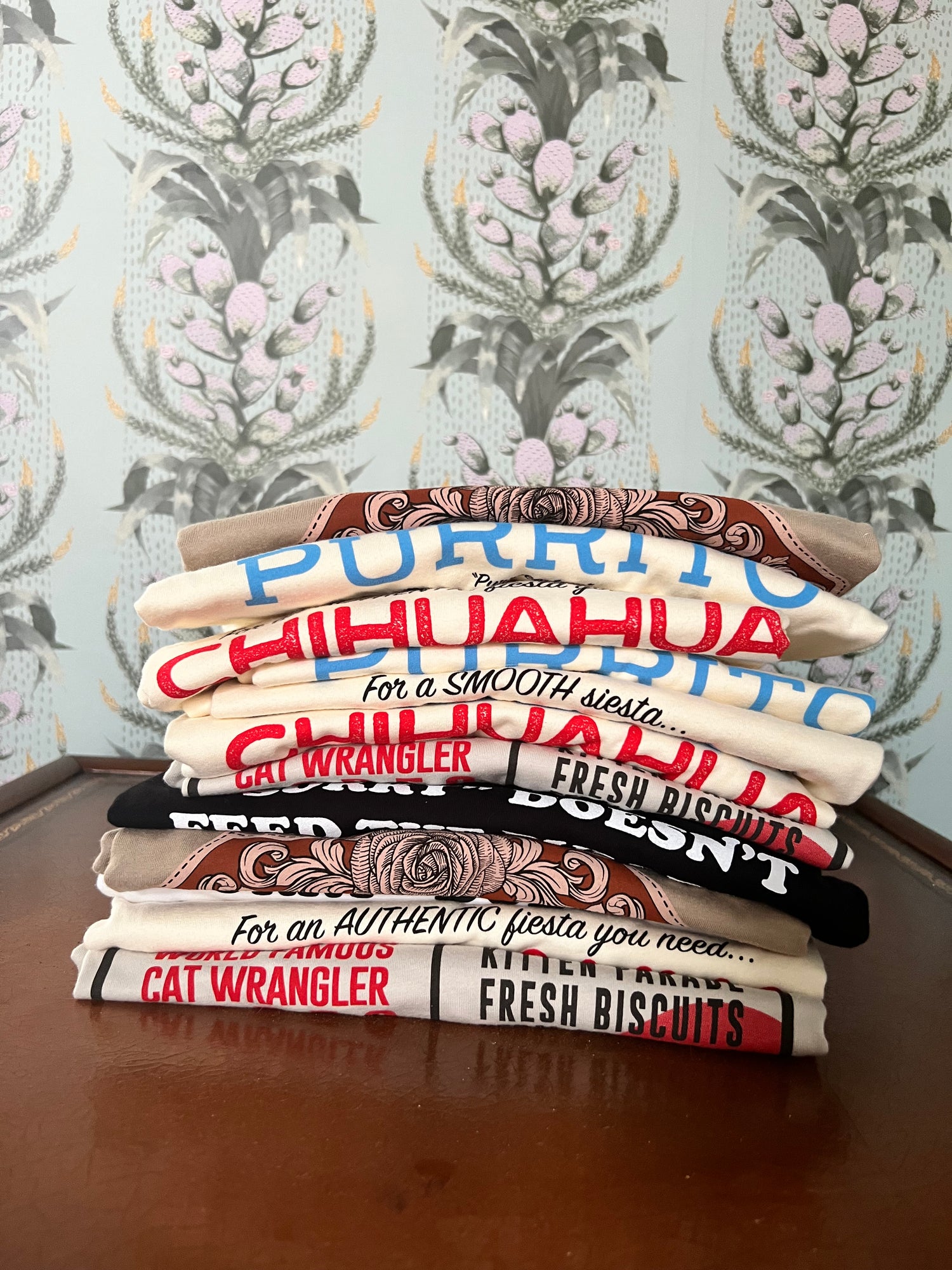 A stack of Pet Rodeo graphic t-shirts in a room with vintage wallpaper