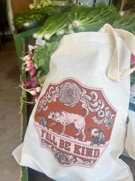 Canvas tote bag with a printed design featuring a dog, pig, and raccoon, and text 'Y'all Be Kind' at a Farm stand.