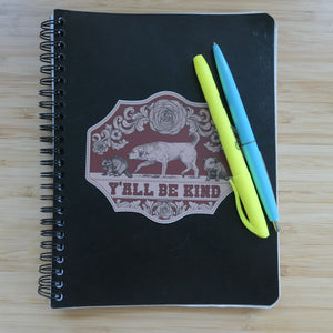 A Black notebook with a sticker that says Y'all Be Kind with illustrations of a dog, raccoon, and piglet